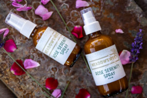 A 2 and 4 oz brown amber bottle lie on a rock strewn with rose petals. The white labels show that the liquid inside is Rose Serenity Spray