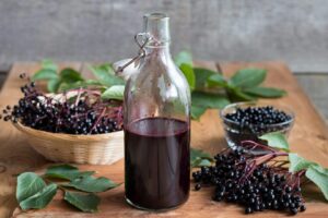 A open hinge top apothocary jug sits with a dark purple Elderberry Syrup on top of a table; surrounded by fresh elderberrie and foliage